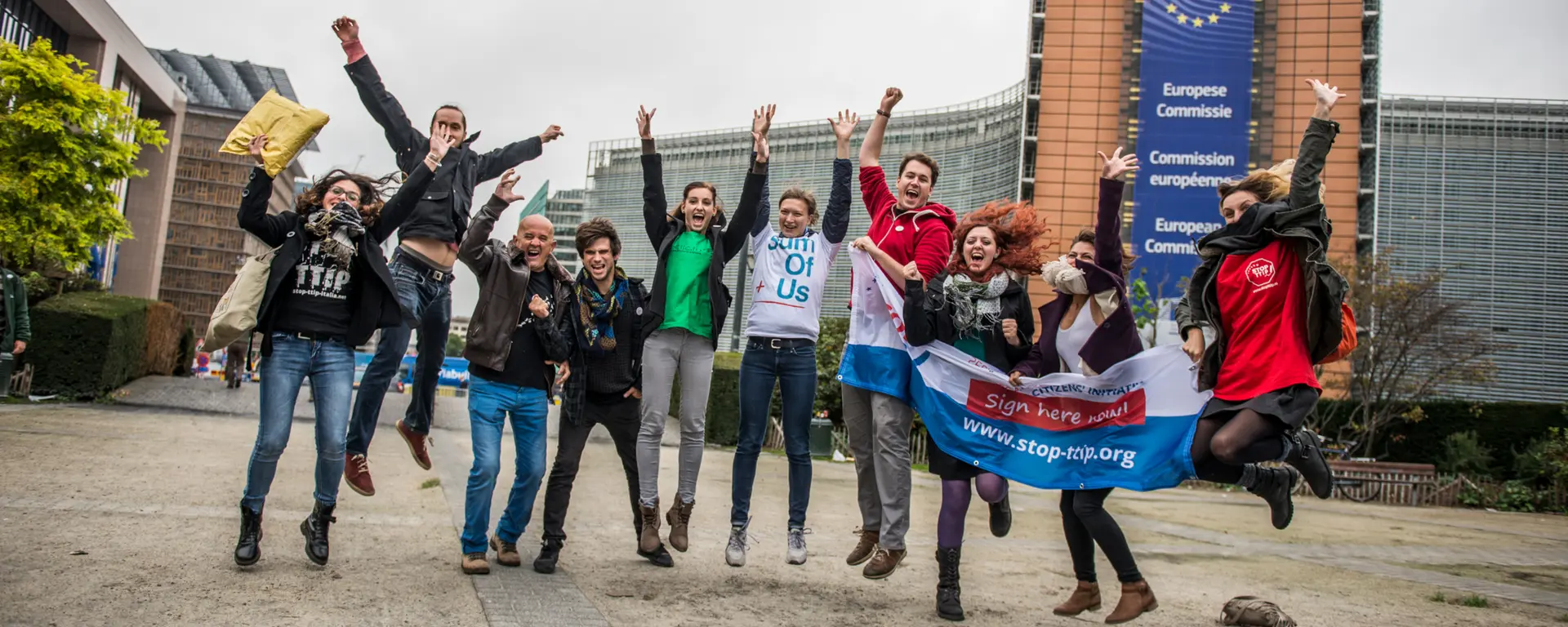 Zu sehen ist ein Gruppenfoto vieler junger Menschen, die hochspringen und sich freuen und ein Transparent hochhalten mit: www.stop-ttip.org.