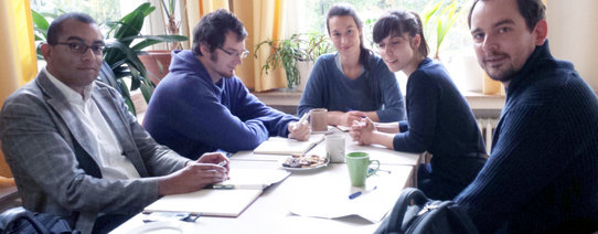 Auf dem Bild sitzen fünf Menschen an einem Tisch und gucken in die Kamera.