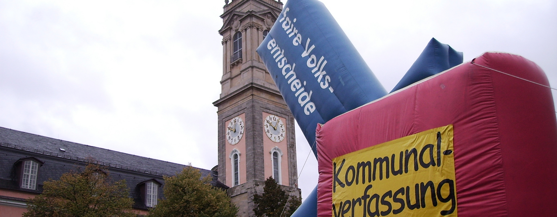Ein circa vier Mal fünf Meter großer, rechteckiges aus Plastik aufgeblasenes Buch, mit der Aufschrift "Kommunal-verfassung" steht vor einer Kirche, mit einem Pfeil aus Plastik darin, welcher die Aufschrift "Faire Volksentscheide" trägt.