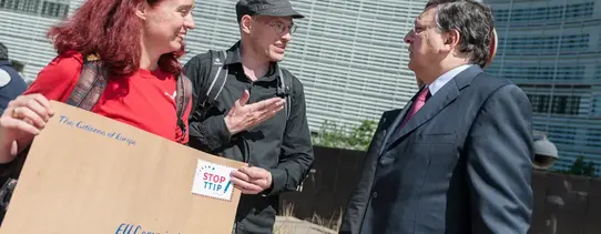 Eine Frau mit einem übergroßen Brief, wo auf der Briefmarke "STOP TTIP" steht, redet mit einem Mann.