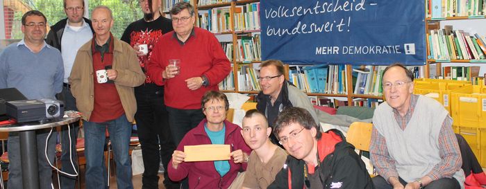 10 Menschen sind vor einem Bücherregal, an welchem ein Transparent angebracht ist, mit dem Text "Volksentscheid-bundesweit!".