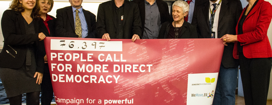 (Unterschriftenübergabe an EU-Parlamentarier | Foto by Edda Dietrich | Lizenz: CC BY-SA 2.0) Neun Menschen halten ein Transparent hoch, welches die Aufschrift hat "PEOPLE CALL FOR MORE DIRECT DEMOCRACY".