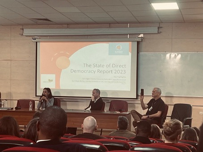 Auf dem Bild ist eine Podiumsdiskussion mit einer Präsentation im Hintergrund an die Wand projiziert.