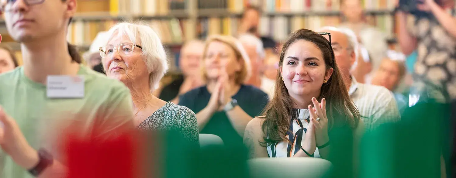 Im Fokus ist eine Frau, in einer sitzenden Menschen Menge, welche klatscht.