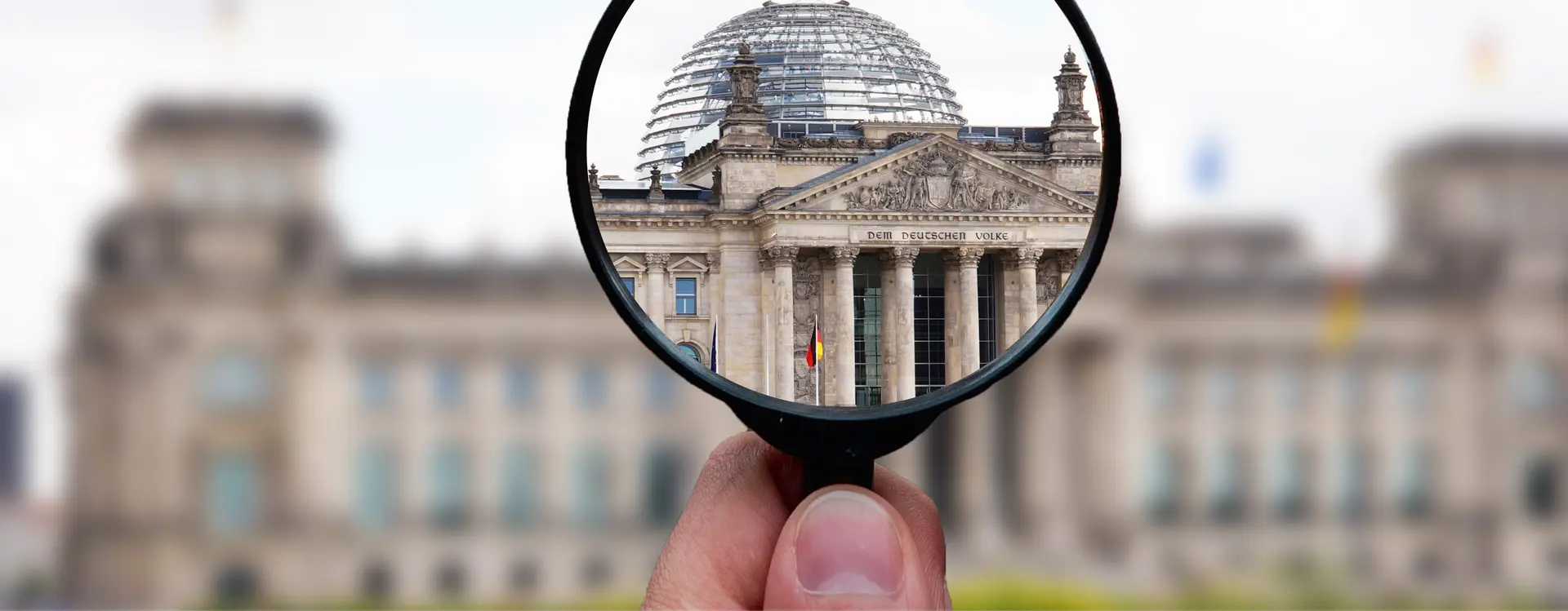 Überschrift: Transparenz. Eine Hand hält eine Lupe auf den Bundestag Eingang auf den Schriftzug "DEM DEUTSCHEN VOLKE". Alles innerhalb der Lupe ist scharf, alles andere ist unscharf (abgesehen von der Hand).