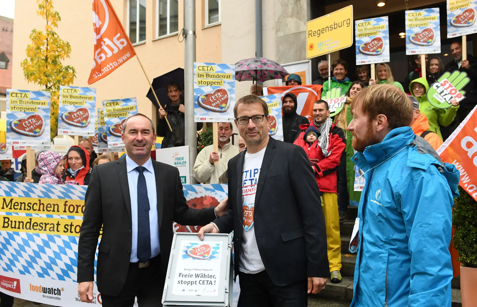 Hubert Aiwanger und Roman Huber gucken in die Kamera und hinter ihnen sind Menschen mit Transparenten.