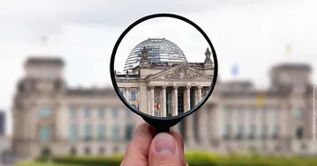 Im Fokus des Bildes ist eine Hand, die eine Lupe auf den Bundestag hält. Alles außerhalb ist verschwommen und der Spruch "DEM DEUTSCHEN VOLK" wird ins Zentrum gerückt.