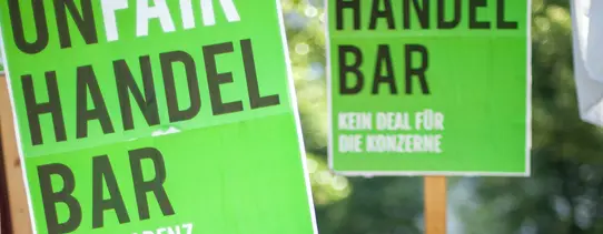 (Fotograf: Jakob Huber/Campact | Lizenz: CC BY-NC 2.0) Auf dem Bild stehen grüne Plakate im Vordergrund, mit der Aufschrift "UNFAIR HANDELBAR", dabei ist "FAIR" weiß und der Rest schwarz. Darunter stehen unterschiedliche Forderungen.