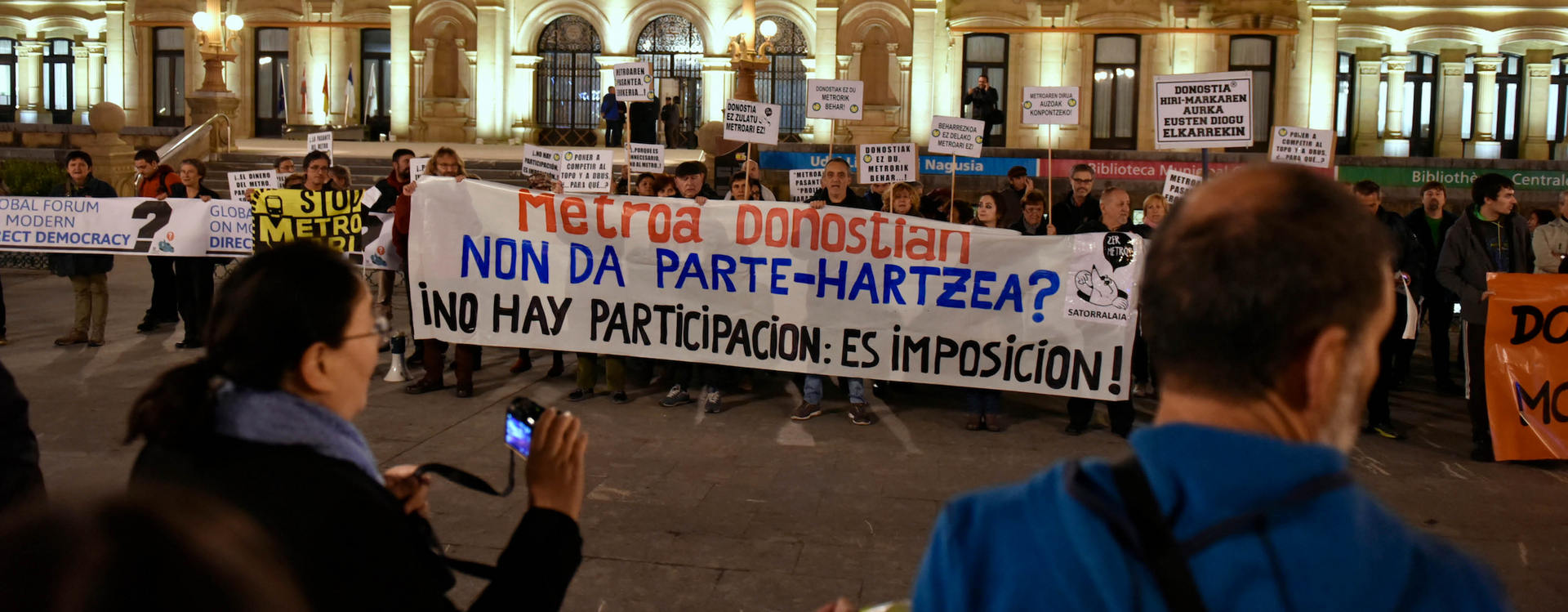Man sieht Menschen die protestieren mit Transparent in ausländischer Schrift, welche vor einem Gebäude stehen.