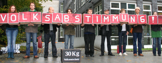 Auf dem Bild sin 9 Menschen, welche Kartons mit Buchstaben hochhalten, welche das Wort "VOLKSABSTIMMUNG!" ergeben.