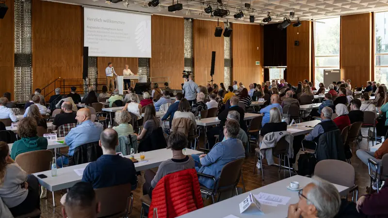 Zahlreiche Frauen und Männer sitzen an langen Tischreihen in einem großen, holzvertäfelten Veranstaltungssaal in Wolfsburg. Sie verfolgen die Begrüßung zum regionalen Dialogforum Nord zur Aufarbeitung der Corona-Pandemie. Auf der Bühne stehen ein Mann mit Mikrofon und eine Frau an einem Rednerpult. Ein Kameramann filmt die Veranstaltung.