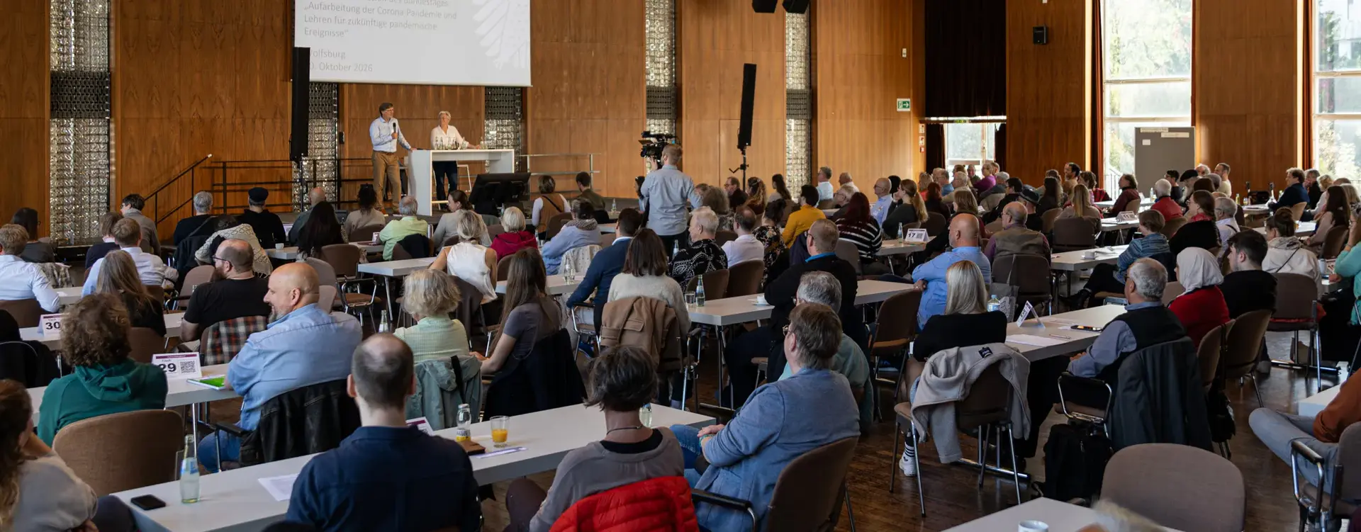 Zahlreiche Frauen und Männer sitzen an langen Tischreihen in einem großen, holzvertäfelten Veranstaltungssaal in Wolfsburg. Sie verfolgen die Begrüßung zum regionalen Dialogforum Nord zur Aufarbeitung der Corona-Pandemie. Auf der Bühne stehen ein Mann mit Mikrofon und eine Frau an einem Rednerpult. Ein Kameramann filmt die Veranstaltung.