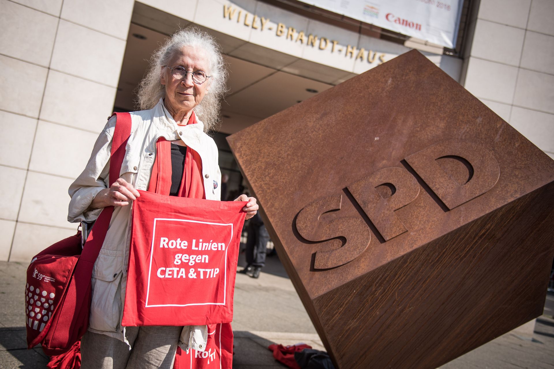 Alter Mensch mit roter Stofftasche, Aufschrift Rote Linien gegen CETA und TTIP. Daneben großer Bronzewürfel mit Relief SPD