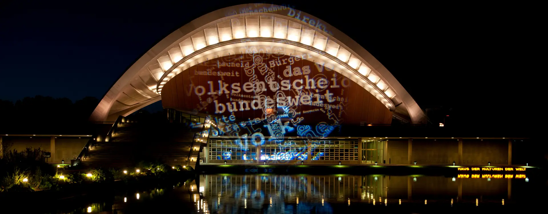 Zu sehen ist das Haus der Kulturen der Welt, ehemals Kongresshalle in Berlin, welches angestrahlt wurde im Zuge einer mobilen Lichtkunstaktion, mit Wörtern wie Volksentscheid, Volk oder Bundesweit.
