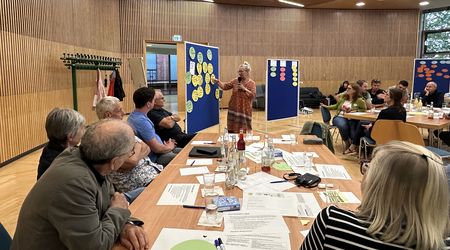 Sitzung des Bürgerrat OPEN_HERZ in Herzberg (Elster): Im Hintergrund erklärt eine Frau die Inhalte von Karten auf einer Pinnwand. Im Vordergrund sitzen Bürgerrat-Mitglieder an einem langen Tisch und einem Tisch rechts auf Höhe der Sprecherin blicken zur Sprecherin und hören ihr zu. 