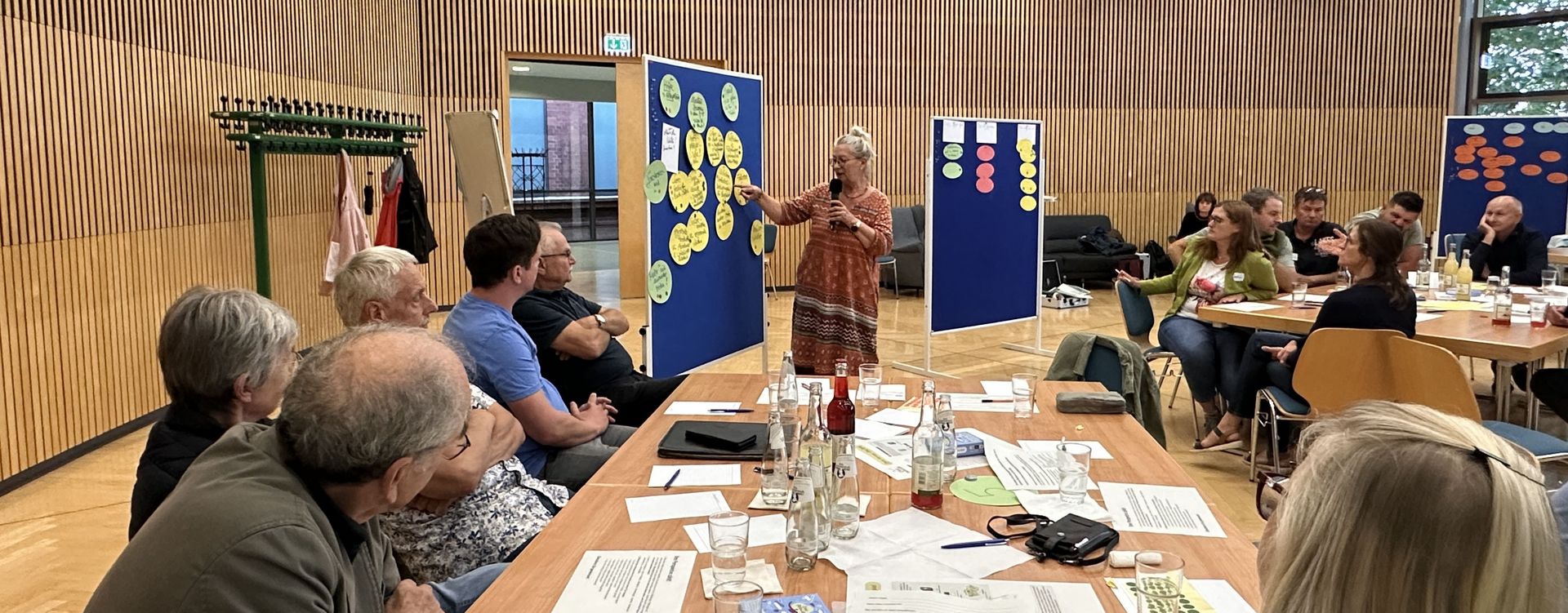 Sitzung des Bürgerrat OPEN_HERZ in Herzberg (Elster): Im Hintergrund erklärt eine Frau die Inhalte von Karten auf einer Pinnwand. Im Vordergrund sitzen Bürgerrat-Mitglieder an einem langen Tisch und einem Tisch rechts auf Höhe der Sprecherin blicken zur Sprecherin und hören ihr zu.