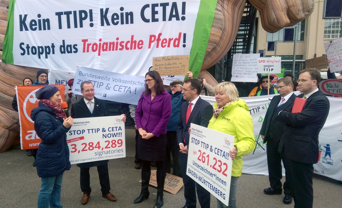 Auf dem Protest ist im Hintergrund ein trojanisches Pferd, mit der Aufschrift auf einem Transparent "Kein TTIP! Kein CETA! Stoppt das Trojanische Pferd!".