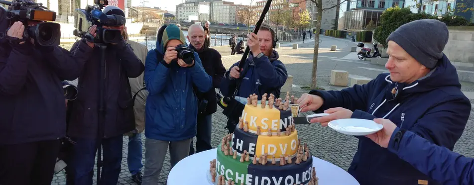 Viele Kameramänner filmen, wie ein Mann aus einer Torte ein Stück Kuchen heraus schneidet, wo Volksentscheid und noch andere Sachen drauf stehen.