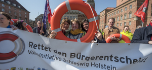 Auf dem Bild ist ein Protest zu sehen. Die Menschen halten ein Transparent hoch, mit dem Text "Rettet den Bürgerentscheid!, Absatz Volksinitiative Schleswig Holstein". Drei Menschen halten dahinter einen Rettungsring hoch und gucken dadurch.