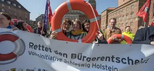 Auf dem Bild ist ein Protest zu sehen. Die Menschen halten ein Transparent hoch, mit dem Text "Rettet den Bürgerentscheid!, Absatz Volksinitiative Schleswig Holstein". Drei Menschen halten dahinter einen Rettungsring hoch und gucken dadurch.