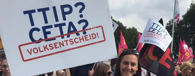 Man sieht einen Protest und im Fokus ist eine Frau, mit dem Text "TTIP? CETA? VOLKSENTSCHEID" auf ein Plakat geschrieben.
