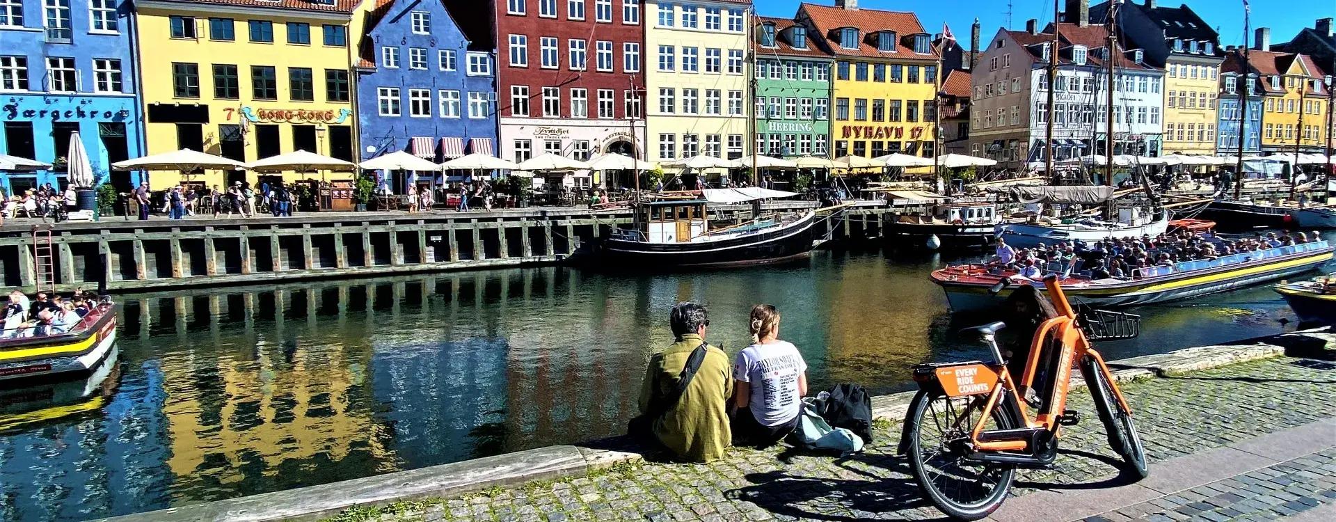 Am Kopenhagener Nyhavn stehen mehrstöckige Häuser mit farbigen Fassaden dicht nebeneinander. Im Kanal liegen zahlreiche Segel- und Motorboote. Am gepflasterten Ufer stehen Fahrräder; im Hintergrund sind weitere Gebäude und Boote zu sehen.