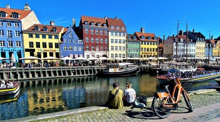 Am Kopenhagener Nyhavn stehen mehrstöckige Häuser mit farbigen Fassaden dicht nebeneinander. Im Kanal liegen zahlreiche Segel- und Motorboote. Am gepflasterten Ufer stehen Fahrräder; im Hintergrund sind weitere Gebäude und Boote zu sehen.