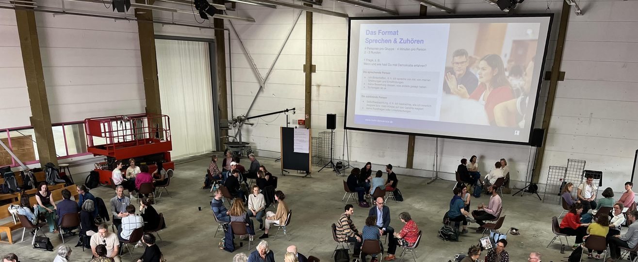 Ein großer Raum mit einer Beamerwand, darin dutzende Menschen die in Vierergruppen sitzen.