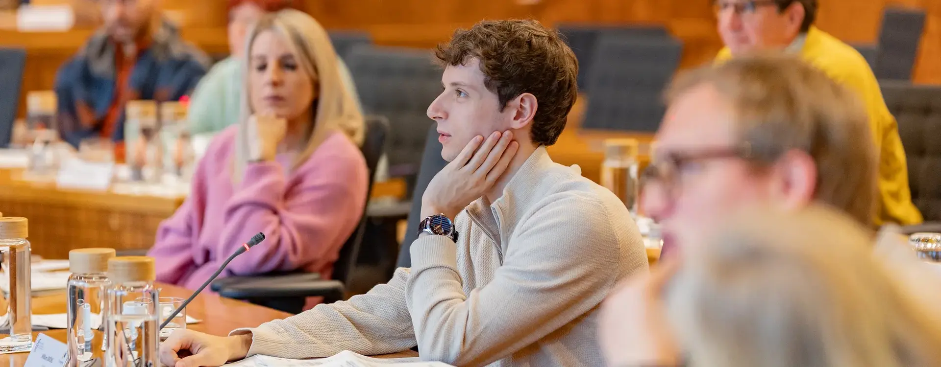 Ein junger Mann mit lockigem braunem Haar und hellem Pullover sitzt an einem Konferenztisch und stützt nachdenklich den Kopf auf seine Hand. Vor ihm liegen Unterlagen, ein Mikrofon und mehrere Wassergläser. Neben und hinter ihm sitzen weitere Frauen und Männer, die aufmerksam zuhören. Der holzvertäfelte Sitzungssaal vermittelt eine sachliche Arbeitsatmosphäre.