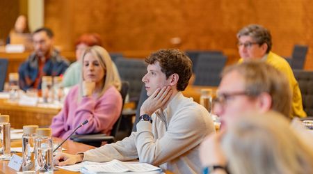 Ein junger Mann mit lockigem braunem Haar und hellem Pullover sitzt an einem Konferenztisch und stützt nachdenklich den Kopf auf seine Hand. Vor ihm liegen Unterlagen, ein Mikrofon und mehrere Wassergläser. Neben und hinter ihm sitzen weitere Frauen und Männer, die aufmerksam zuhören. Der holzvertäfelte Sitzungssaal vermittelt eine sachliche Arbeitsatmosphäre.