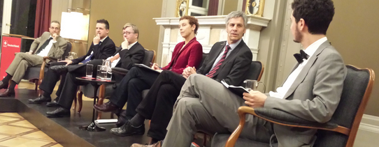 Sechs Menschen sitzen in Anzügen auf hochwertigen Stühlen in einem schönen Saal.