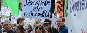 Auf dem Bild ist ein Plakat bei einem Protest im Fokus, mit dem Text "Demokratie jetzt auch in der EU".