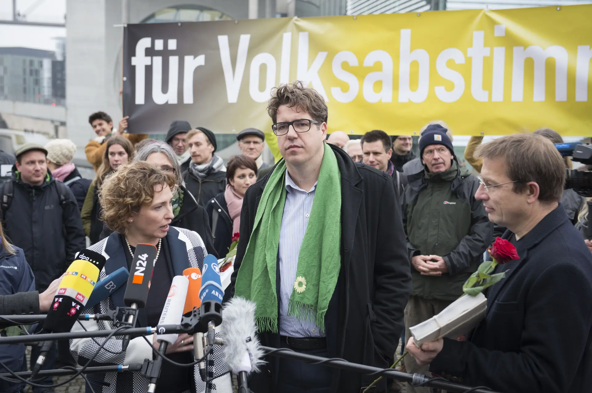 Vor einem Protest stehen drei Menschen vor Mikrofonen.