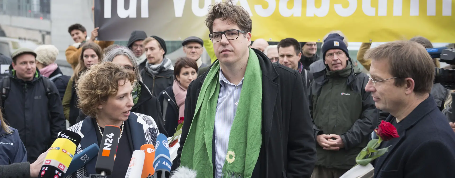 Drei Menschen stehen vor Mikrofonen und Kameras und dahinter stehen Menschen mit einem Transparent, mit der Aufschrift "für Volksabstimmungen".