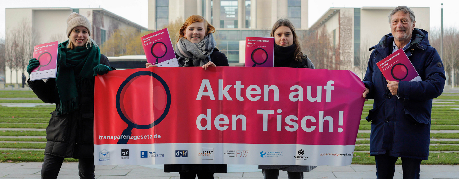 Es sind vier Menschen zu sehen, die ein Transparent hochhalten, wo man eine Lupe und darunter transparenzgesetz.de lesen kann. Rechts davon steht groß: Akten auf den Tisch!. In den unteren 10% des Transparents kann man die Beteiligten Organisationen ablesen, wie "Frag den Staat", oder "Mehr Demokratie".