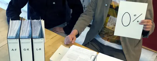 (Dr. Michael Efler & Charlie Rutz beim Vorbereiten der Verfassungsbeschwerde; Foto-Verwendung: Mehr Demokratie e.V. | Lizenz: CC BY-NC 2.0) Zwei Männer sitzen an einem Tisch vor Aktenordnen und einer hält ein Blatt Papier hoch, wo "0%" drauf steht.