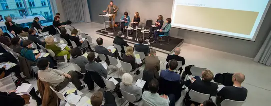 Ein Vortrag von fünf Menschen ist zu sehen, und circa 40 Personen, welche sitzend zuhören.