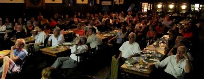 In einem Saal sitzen circa 100 Menschen auf Stühlen und gucken nach vorne.
