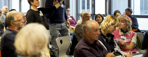 Eine Frau hält einem Mann das Mikrofon hin (auf einer Veranstaltung), damit er etwas sagen oder fragen kann, während der Rest auf Stühlen sitzt und zuhört.