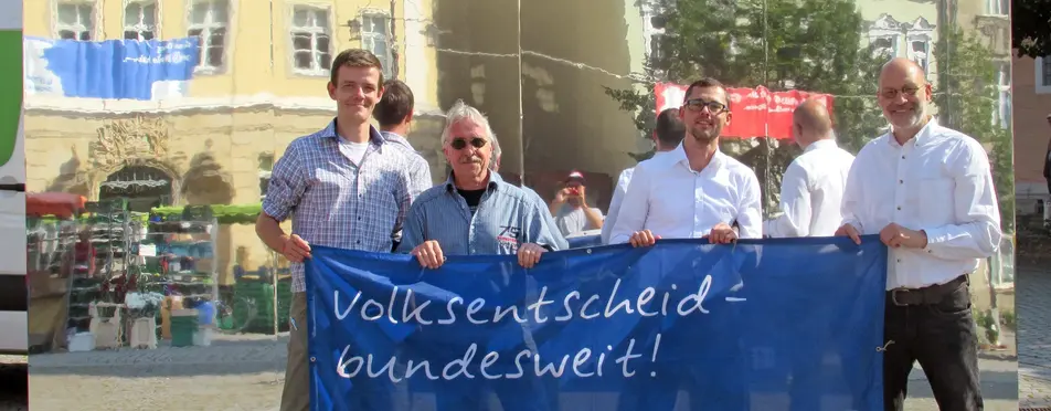 Vier Menschen stehen vor einem Spiegel, mit der Überschrift "...wer ist der Souverän?" und halten zudem ein Transparent hoch, mit der Aufschrift "Volksentscheid - bundesweit!".
