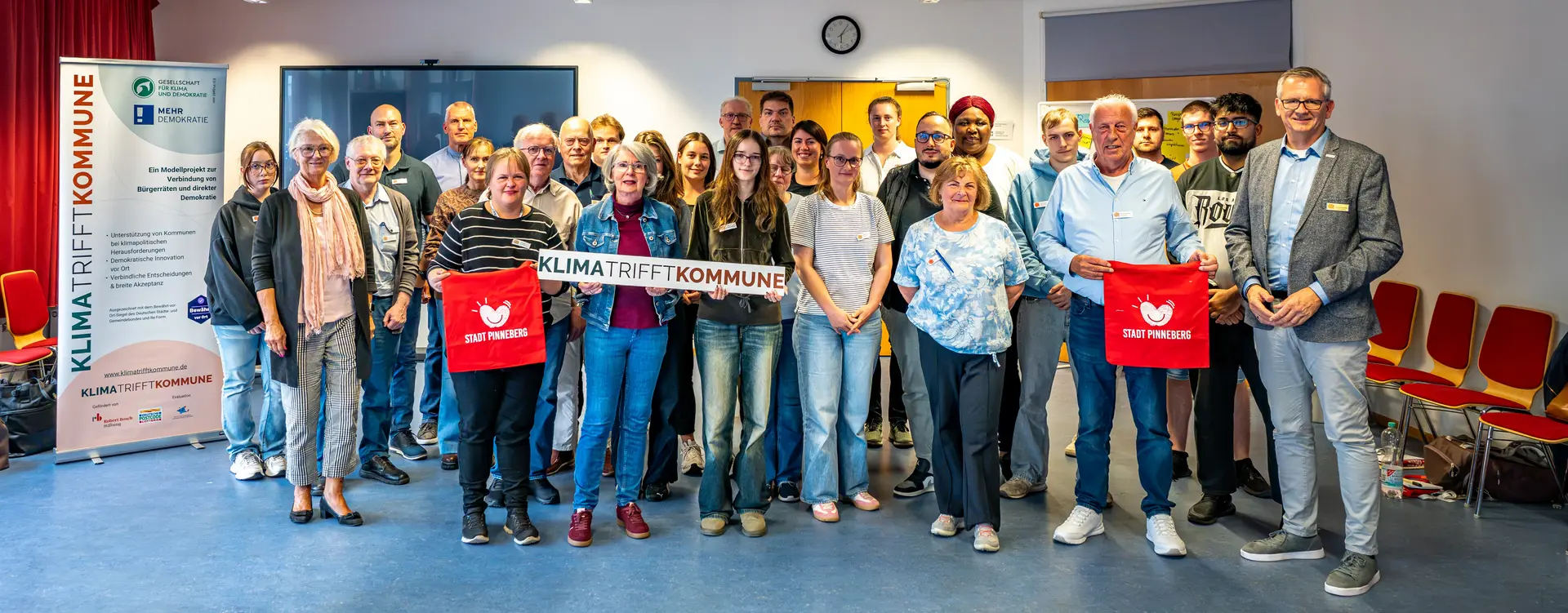 Eine größere Gruppe von Frauen und Männern unterschiedlichen Alters steht für ein Gruppenfoto in einem hellen Veranstaltungsraum. Die Personen stehen in mehreren Reihen dicht beieinander und blicken überwiegend lächelnd in die Kamera. In der Mitte halten zwei Frauen ein weißes Banner mit der Aufschrift „Klima trifft Kommune“. Zwei Personen in der vorderen Reihe halten rote Stofftaschen der Stadt Pinneberg. Links neben der Gruppe steht ein großes Roll-up des Projekts „Klima trifft Kommune“. Im Hintergrund sind eine Leinwand, eine Uhr und Teile der Veranstaltungstechnik zu sehen. Rechts stehen mehrere rote Stühle an der Wand.