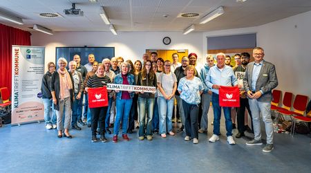 Eine größere Gruppe von Frauen und Männern unterschiedlichen Alters steht für ein Gruppenfoto in einem hellen Veranstaltungsraum. Die Personen stehen in mehreren Reihen dicht beieinander und blicken überwiegend lächelnd in die Kamera. In der Mitte halten zwei Frauen ein weißes Banner mit der Aufschrift „Klima trifft Kommune“. Zwei Personen in der vorderen Reihe halten rote Stofftaschen der Stadt Pinneberg. Links neben der Gruppe steht ein großes Roll-up des Projekts „Klima trifft Kommune“. Im Hintergrund sind eine Leinwand, eine Uhr und Teile der Veranstaltungstechnik zu sehen. Rechts stehen mehrere rote Stühle an der Wand.