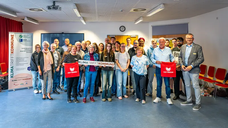 Eine größere Gruppe von Frauen und Männern unterschiedlichen Alters steht für ein Gruppenfoto in einem hellen Veranstaltungsraum. Die Personen stehen in mehreren Reihen dicht beieinander und blicken überwiegend lächelnd in die Kamera. In der Mitte halten zwei Frauen ein weißes Banner mit der Aufschrift „Klima trifft Kommune“. Zwei Personen in der vorderen Reihe halten rote Stofftaschen der Stadt Pinneberg. Links neben der Gruppe steht ein großes Roll-up des Projekts „Klima trifft Kommune“. Im Hintergrund sind eine Leinwand, eine Uhr und Teile der Veranstaltungstechnik zu sehen. Rechts stehen mehrere rote Stühle an der Wand.
