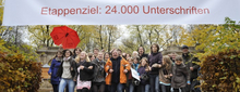 Auf dem Foto stehen circa 20 Personen zwischen zwei Hecken und darüber ist ein Banner, mit der Aufschrift "Schule in Freiheit, Absatz Etappenziel: 24.000 Unterschriften".