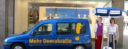 demokratiemobil Ein blaues Auto, wo "Mehr Demokratie" drauf steht, hat rechts neben sich einen Infostand von Mehr Demokratie auf dem Bild.