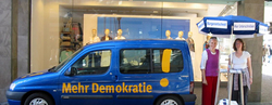 demokratiemobil Ein blaues Auto, wo "Mehr Demokratie" drauf steht, hat rechts neben sich einen Infostand von Mehr Demokratie auf dem Bild.