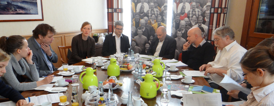 Auf dem Bild sind 10 Menschen mit vielen Papieren und unterhalten sich.