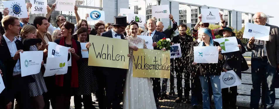Auf dem Bild ist eine Braut mit einem Plakat (Volksentscheid) und den Bräutigam ebenfalls mit einem Plakat (Wahlen). Im Hintergrund stehen Menschen ebenfalls mit Plakaten.