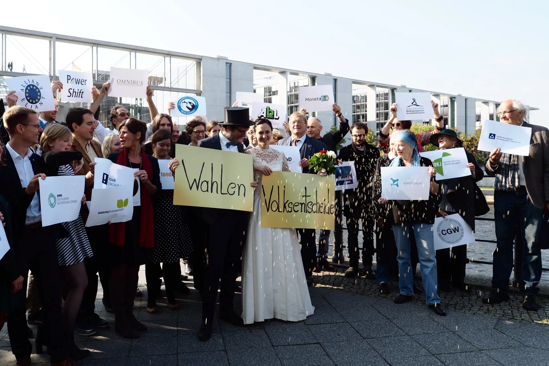 Eine Braut und ihr Bräutigam halten Plakate hoch, mit den Worten "Wahlen" und "Volksentscheide".