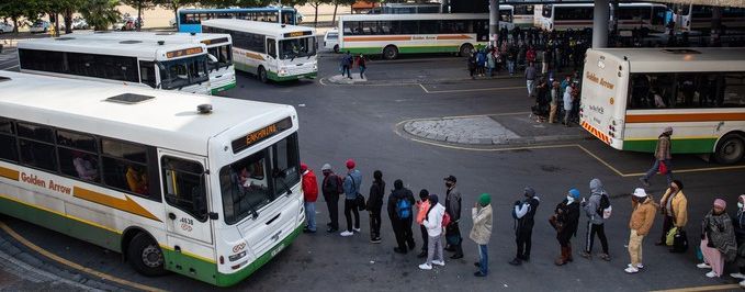 Blick über einen großen Busbahnhof in Kapstadt. Mehrere Busse stehen nebeneinander an den Haltestellen. Zwischen den Fahrzeugen und entlang der überdachten Bahnsteige sind zahlreiche Menschen unterwegs oder warten auf ihre Abfahrt. Im Hintergrund erstreckt sich die dichte Bebauung der Stadt; dahinter erhebt sich die Berglandschaft Kapstadts. 